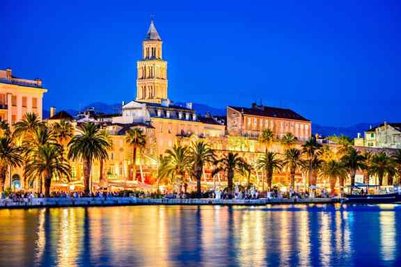 Photo of waterfront in Split at night, all lit up. With palm trees and lots of people and reflections in the water.